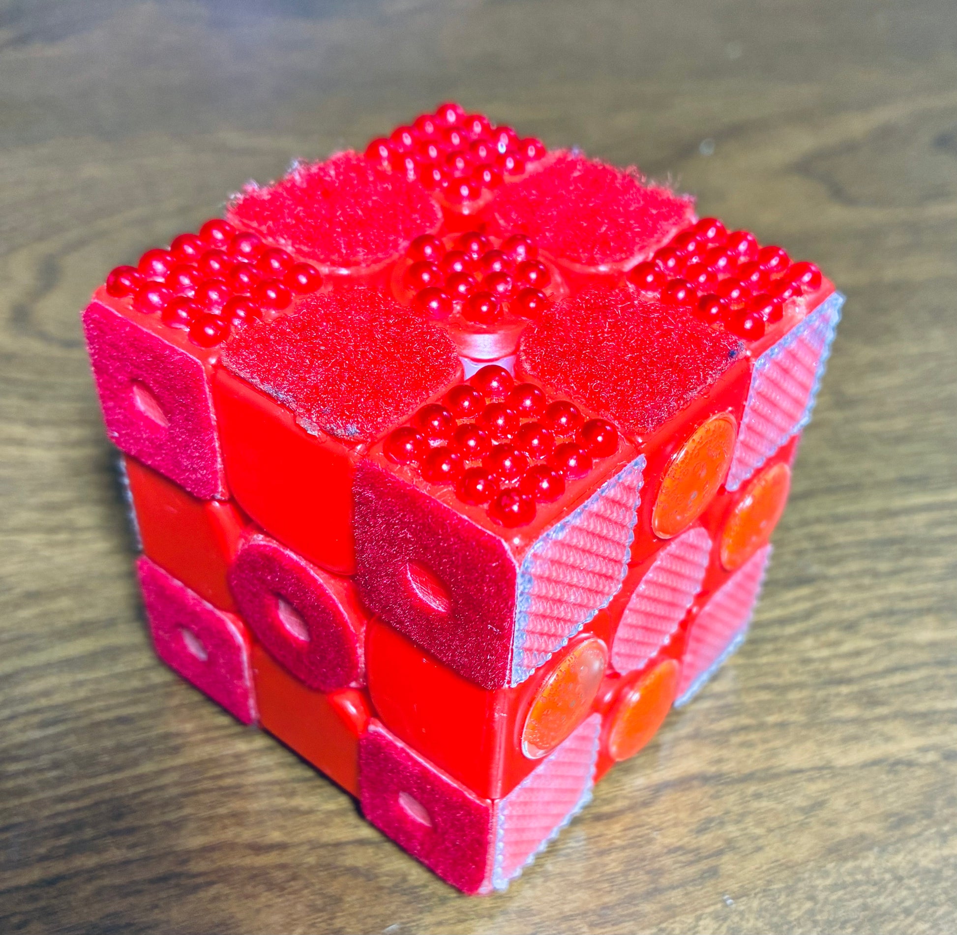 Texture Cube textured Rubik's cube pictured in checkerboard pattern to show off six distinct sides, which are all red. One side is smooth plastic, one has a large, round piece on each square, one has soft velvet with a punched hole in the centers, one has a rough, grippy texture, one has a fluffy, carpet-like Velcro texture, and one is a bumpy texture made of pearls.